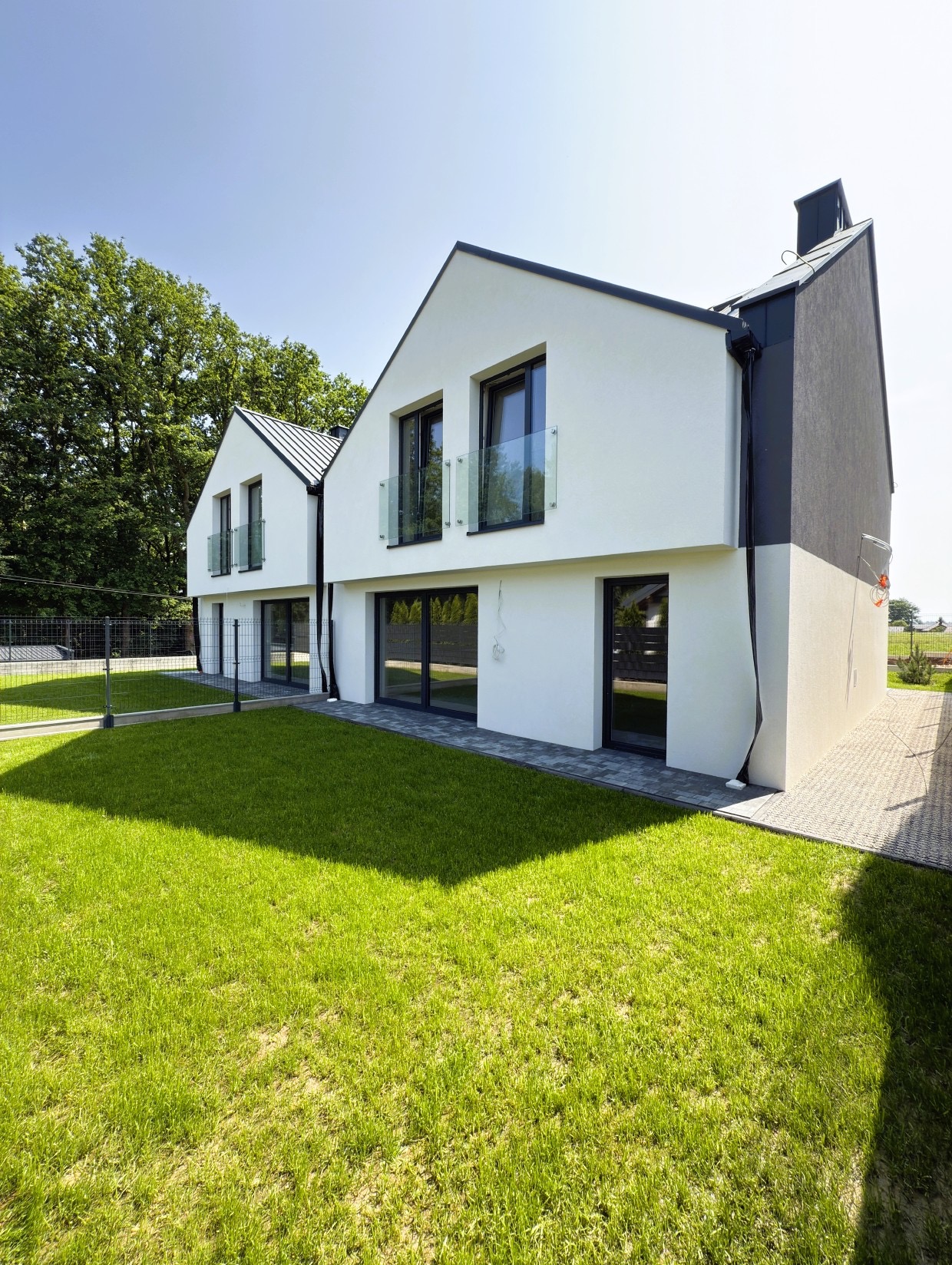 Nowoczesny, dwukondygnacyjny dom z białymi ścianami i szarym akcentem, z balkonami z przezroczystą balustradą i dużymi oknami, otoczony zielonym trawnikiem.