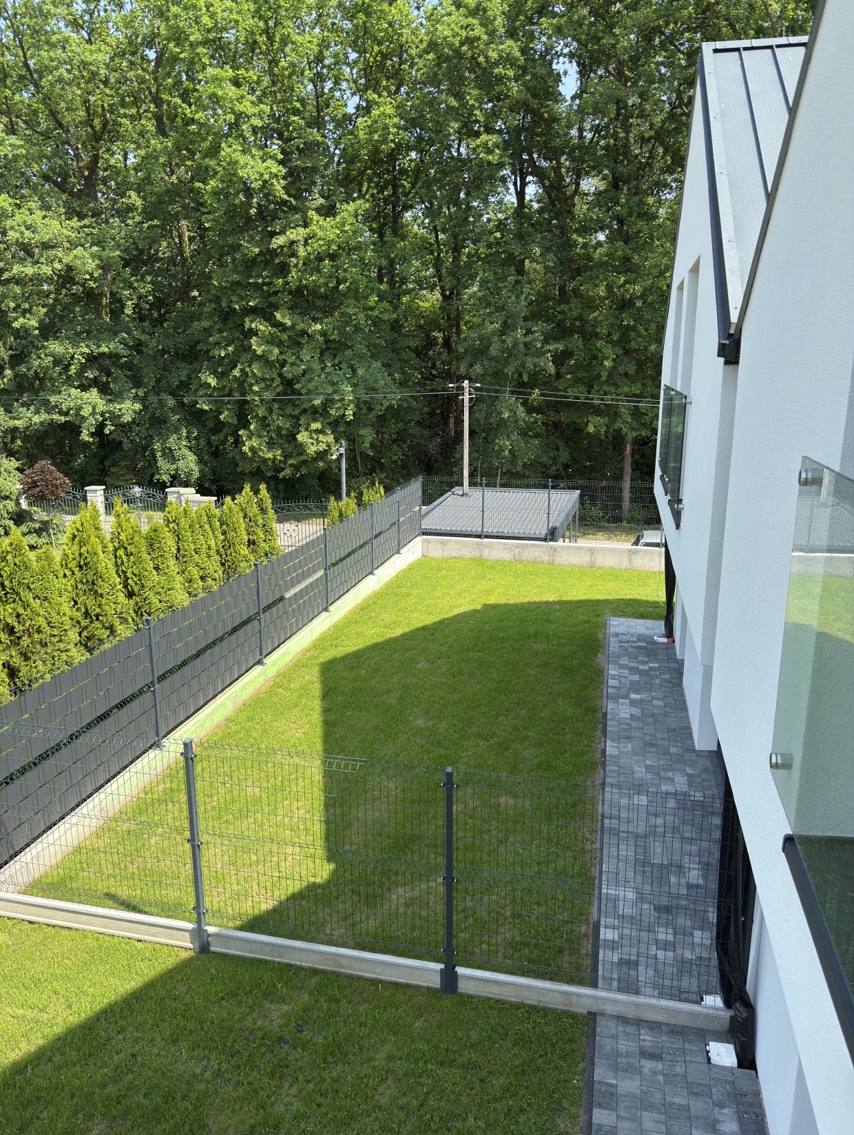 Nowoczesny dom z ogrodem: widok z góry na zielony trawnik, szary płot z panelami, ścieżkę z kostki i fragment białej elewacji. Krzyszkowice LAWENDOWE DOMY.