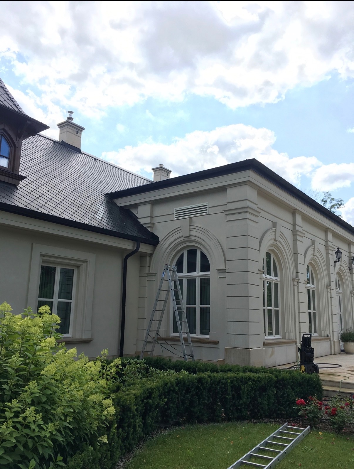 Elewacja dworku w Krzyszkowicach z jasną fasadą, łukowymi oknami i drabiną opartą o ścianę. W tle błękitne niebo z chmurami. Widoczny fragment dachu i zieleni.