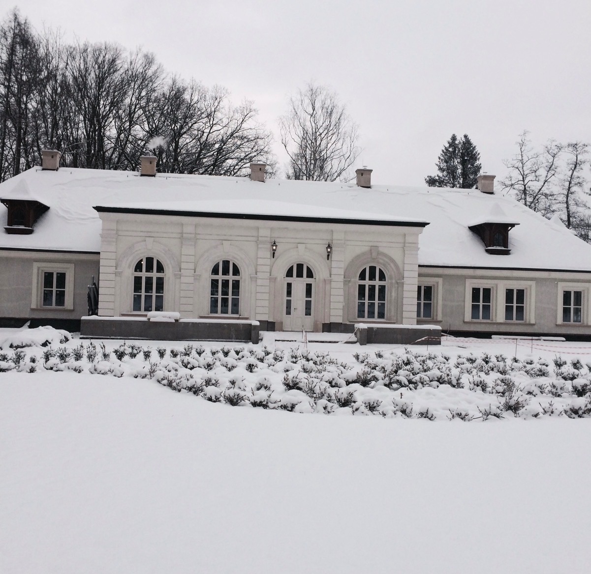 Dwór w zimowej scenerii, pokryty śniegiem, z widoczną architekturą klasycystyczną i oknami łukowymi. Śnieżny krajobraz podkreśla biel budynku.