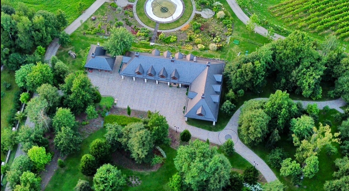 Widok z lotu ptaka na dwór w Krzyszkowicach otoczony zielenią, z fontanną i ścieżkami. Uwagę zwraca symetryczny układ budynku i starannie zaprojektowany ogród.