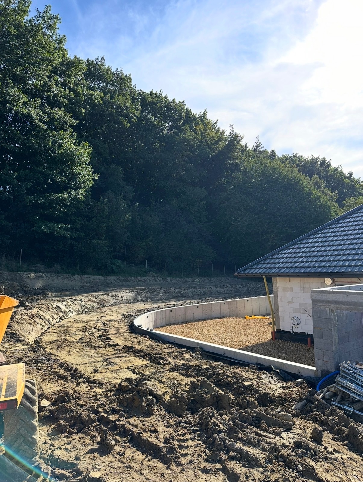 Niwelacja terenu pod budowę domu z murem oporowym w kształcie łuku. Widoczny fragment budynku w stanie surowym otwartym i żółta koparka. Tło: las i błękitne niebo.