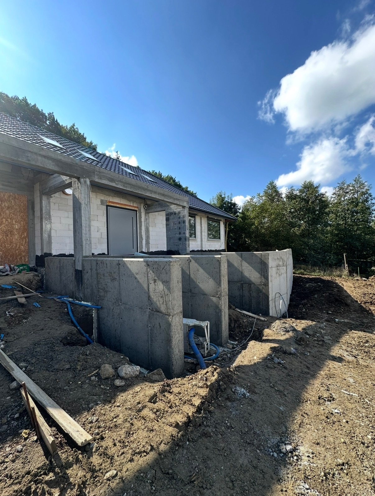 Budowa domu w trakcie: wzniesione fundamenty z betonu, ściany z bloczków, widoczne niebieskie rury odwodnienia, niwelacja terenu. Słoneczny dzień.