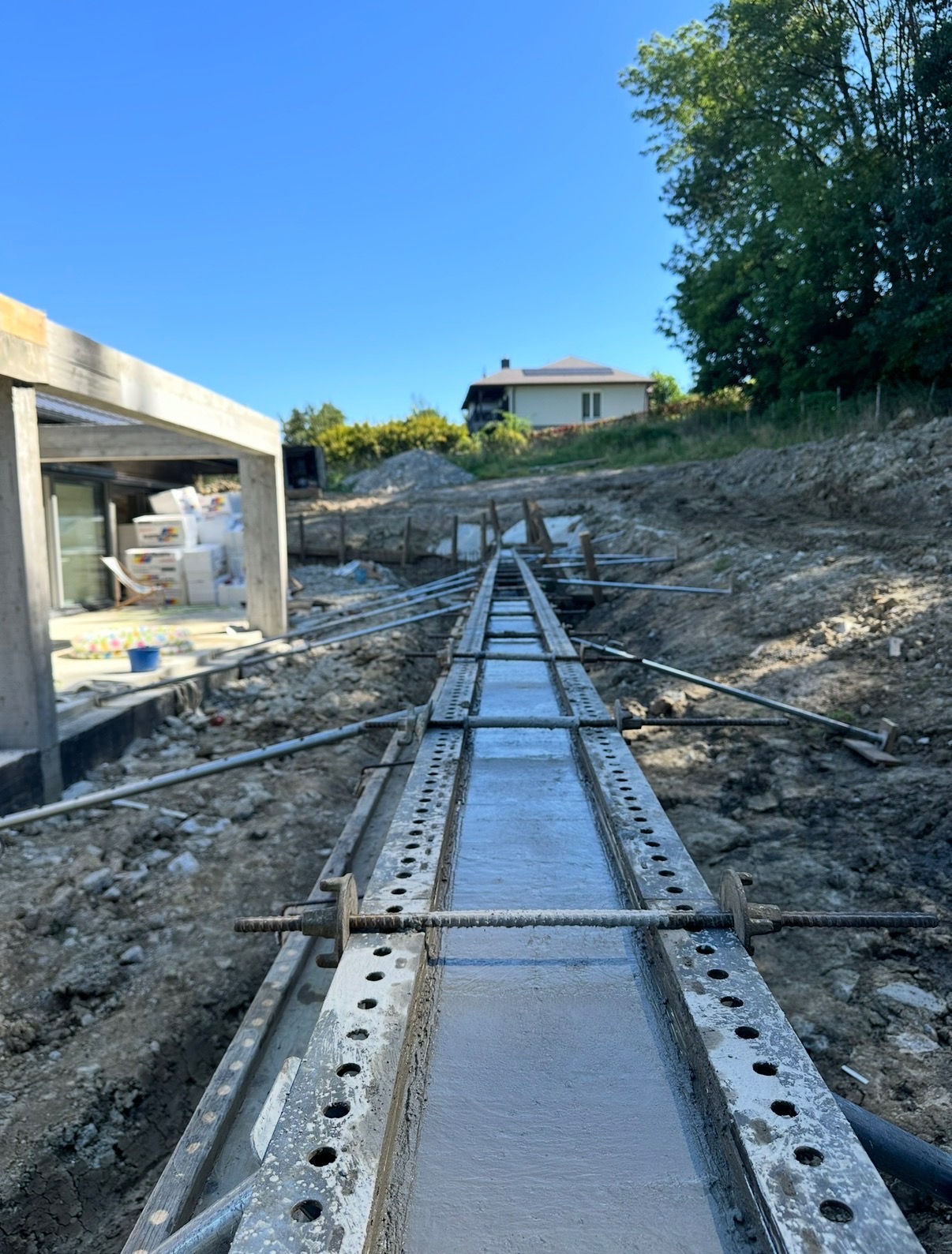 Wąski, zalany betonem szalunek muru oporowego, widoczny z perspektywy ziemi, z fragmentem budynku w tle i błękitnym niebem. Prace ziemne w toku.