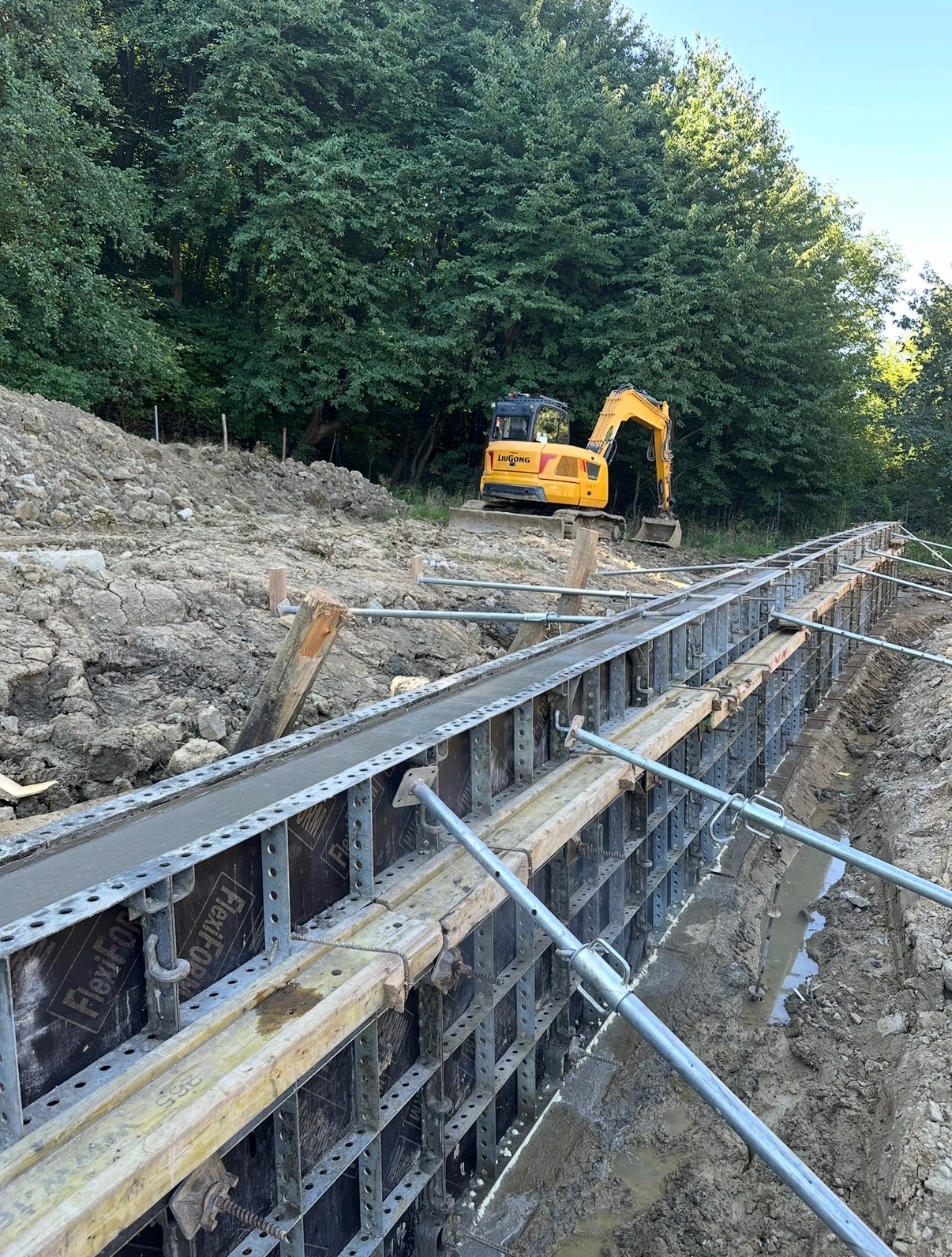 Wykop z szalunkiem wypełnionym betonem, w tle żółta koparka Liugong i drzewa. Widoczne metalowe podpory szalunku. Prace ziemne i betonowanie w toku.