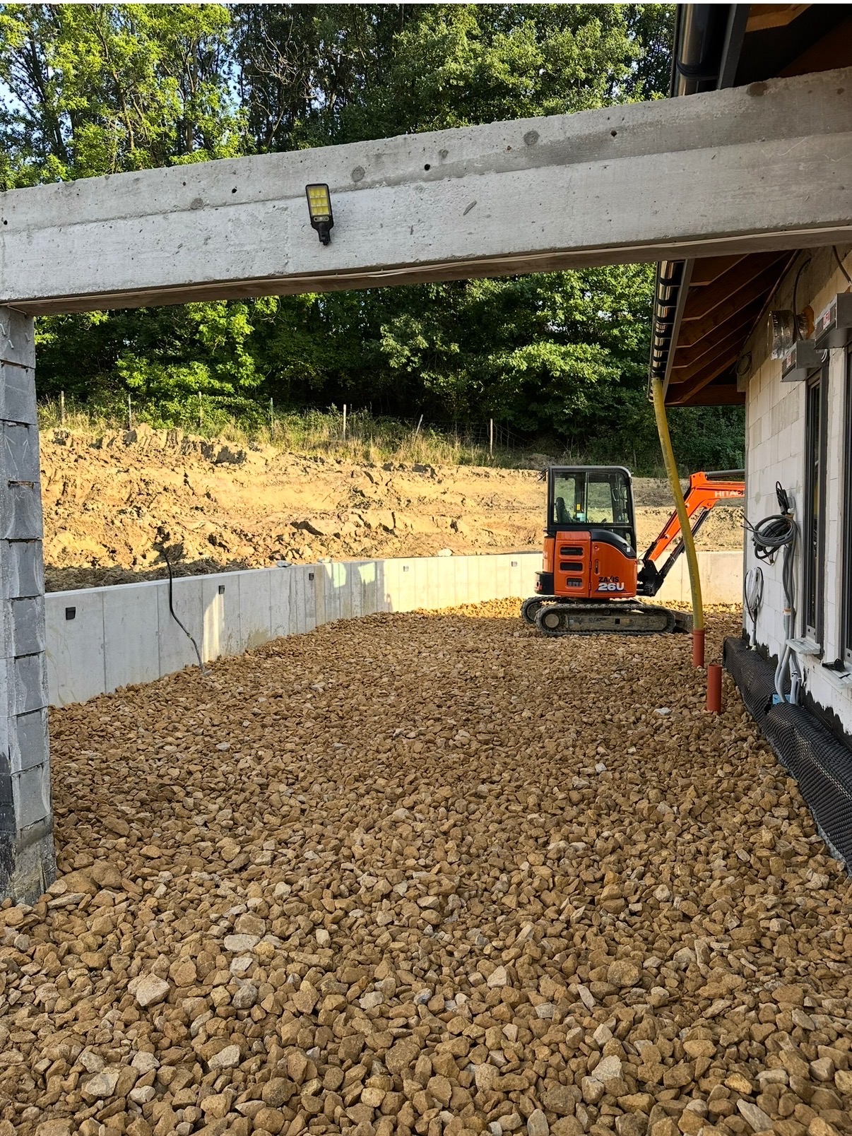 Teren po niwelacji z murem oporowym i odwodnieniem, widoczna mała koparka Hitachi 26U, podsypka z kamienia, fragment budynku z izolacją pionową.