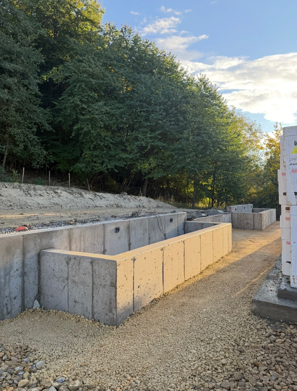 Wykop pod fundamenty z betonowymi ścianami oporowymi na żwirowym podłożu, w tle zieleń drzew i błękitne niebo z chmurami. Widoczne bloczki styropianowe.