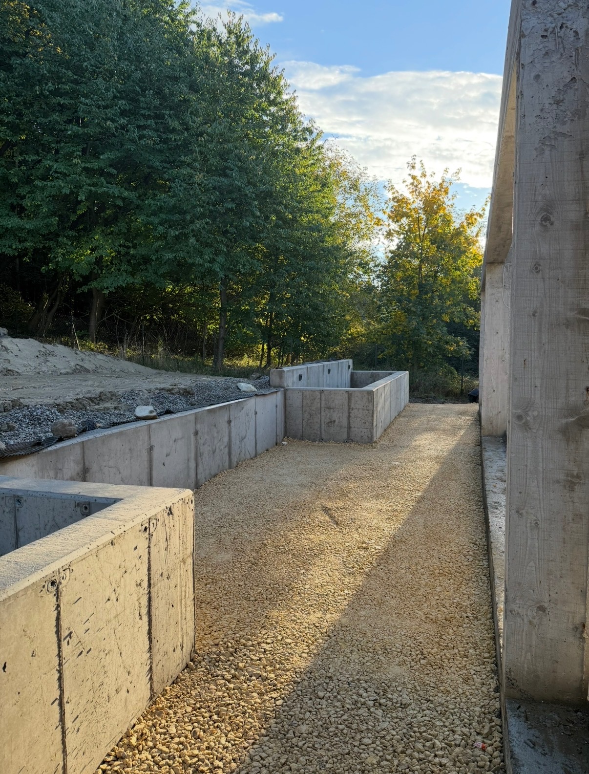 Betonowe mury oporowe w wykopie, wypełnione żwirem, z widocznym fragmentem konstrukcji budynku w tle i zielenią drzew. Ujęcie z poziomu gruntu, ciepłe światło.