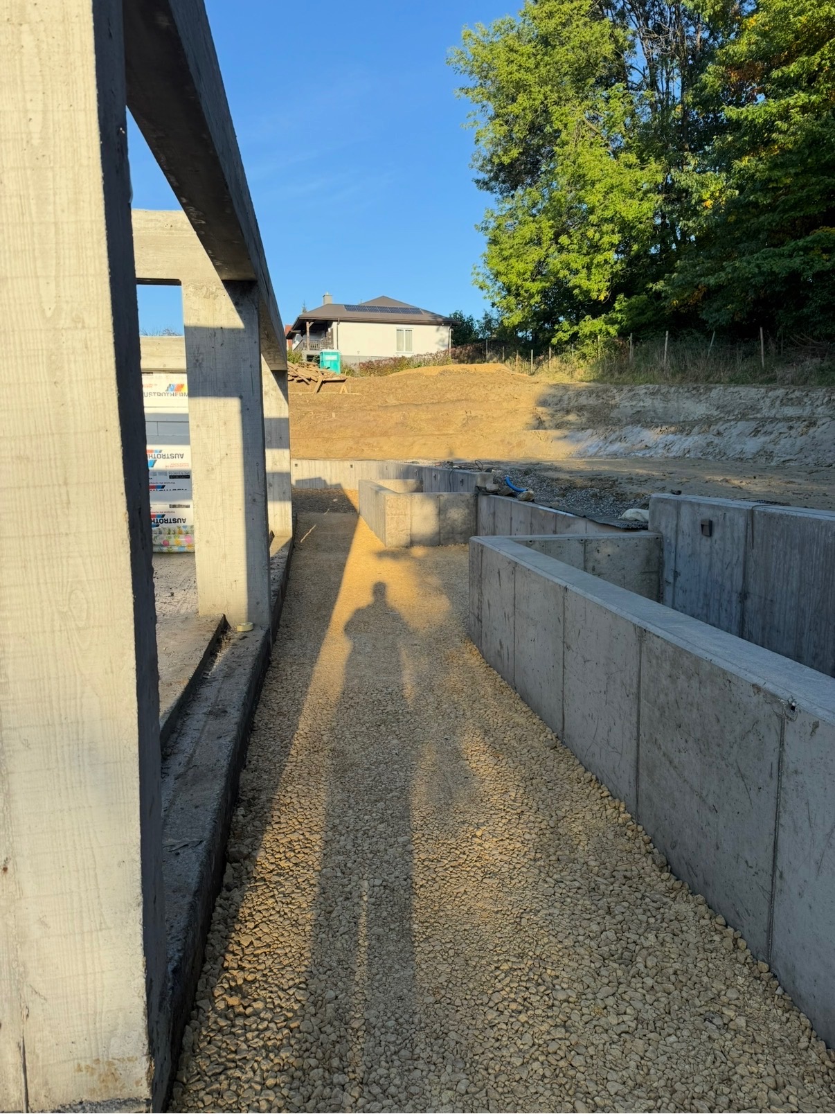 Betonowe mury oporowe w trakcie budowy, widoczny cień osoby, teren po niwelacji, w tle dom z panelami słonecznymi, błękitne niebo.