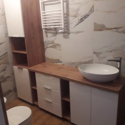 J.J.Meble - Nowoczesna łazienka z umywalką nablatową, szafkami z białymi frontami i drewnianym blatem, wiszącym grzejnikiem drabinkowym na ścianie z marmurowym wzorem.