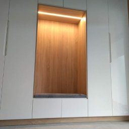 J.J.Meble - Biała szafa wnękowa z drewnianym wnęką z podświetleniem LED i szarą poduszką na siedzisku.