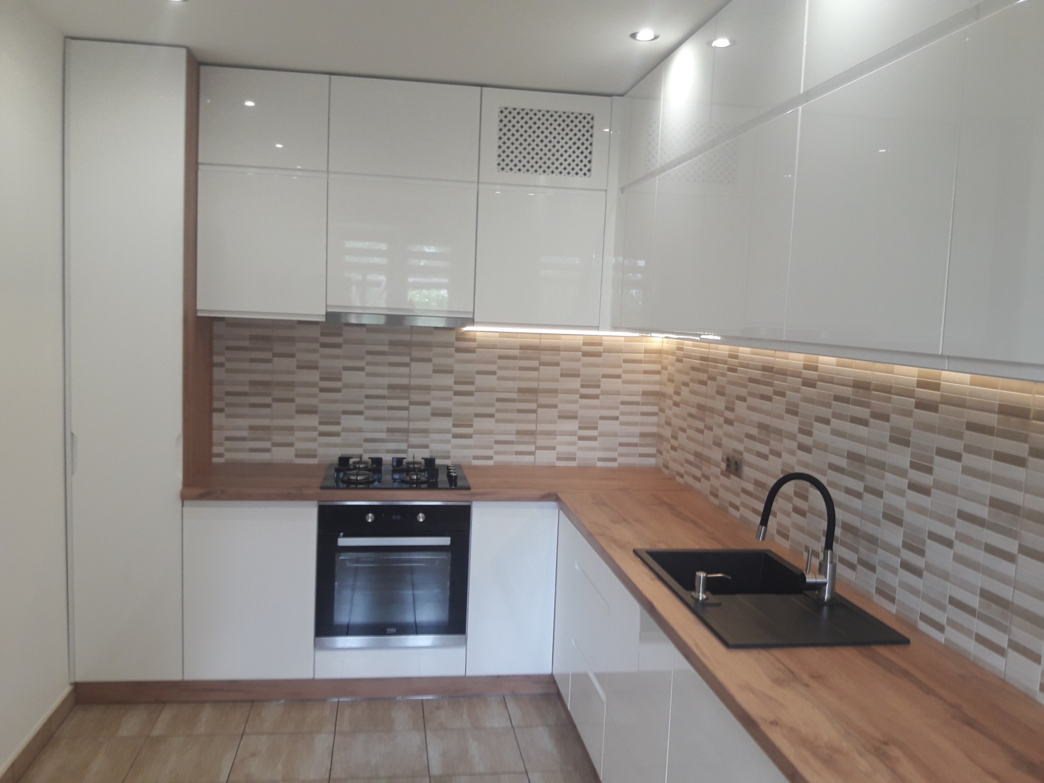 Biała, nowoczesna kuchnia z drewnianym blatem, płytkami w odcieniach beżu i brązu nad blatem oraz czarnym zlewem i baterią.