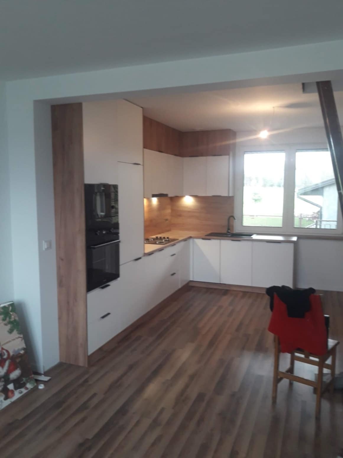 Nowoczesna kuchnia z białymi frontami i drewnianym blatem, widoczna z perspektywy podłogi, z krzesłem z ubraniami w tle i niedokończonym elementem konstrukcyjnym przy oknie.