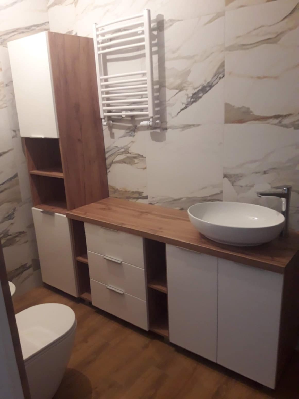 Nowoczesna łazienka z umywalką nablatową, szafkami z białymi frontami i drewnianym blatem, wiszącym grzejnikiem drabinkowym na ścianie z marmurowym wzorem.