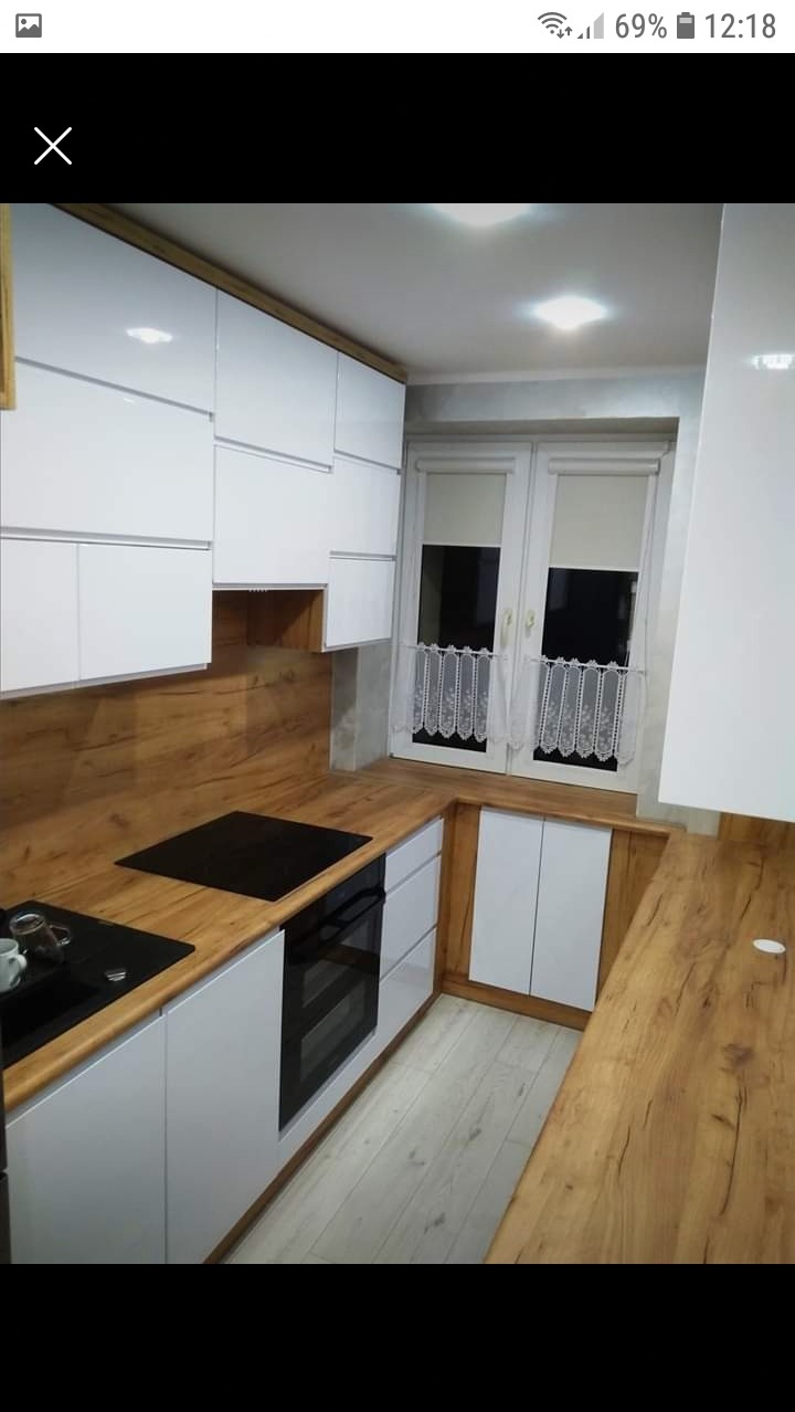 Nowoczesna kuchnia z białymi szafkami i drewnianym blatem, płyta indukcyjna i piekarnik w zabudowie, okno z koronkową firanką.