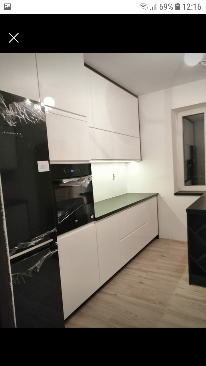 Nowoczesna kuchnia z białymi szafkami i czarnym blatem, sprzęt AGD jeszcze w folii ochronnej, podświetlenie LED pod szafkami wiszącymi, drewniana podłoga.