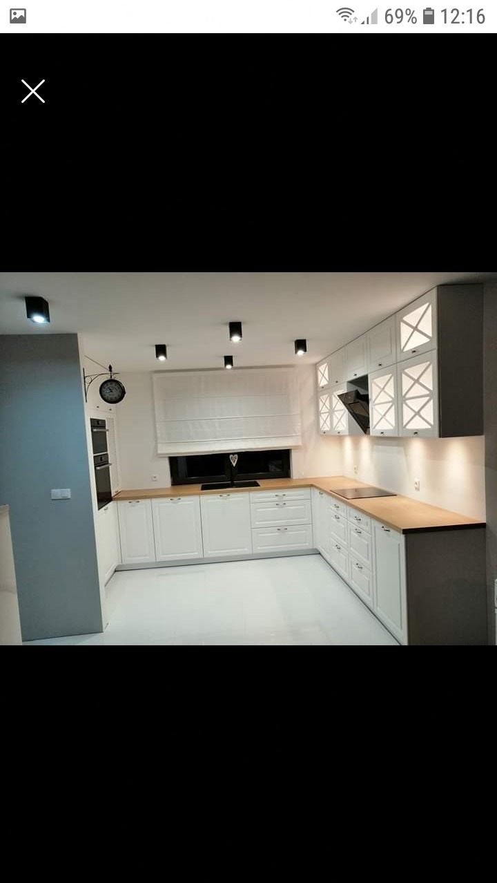 Jasna kuchnia w stylu skandynawskim z białymi szafkami z drewnianymi blatami, oświetleniem punktowym i dekoracyjnymi elementami w górnych szafkach.