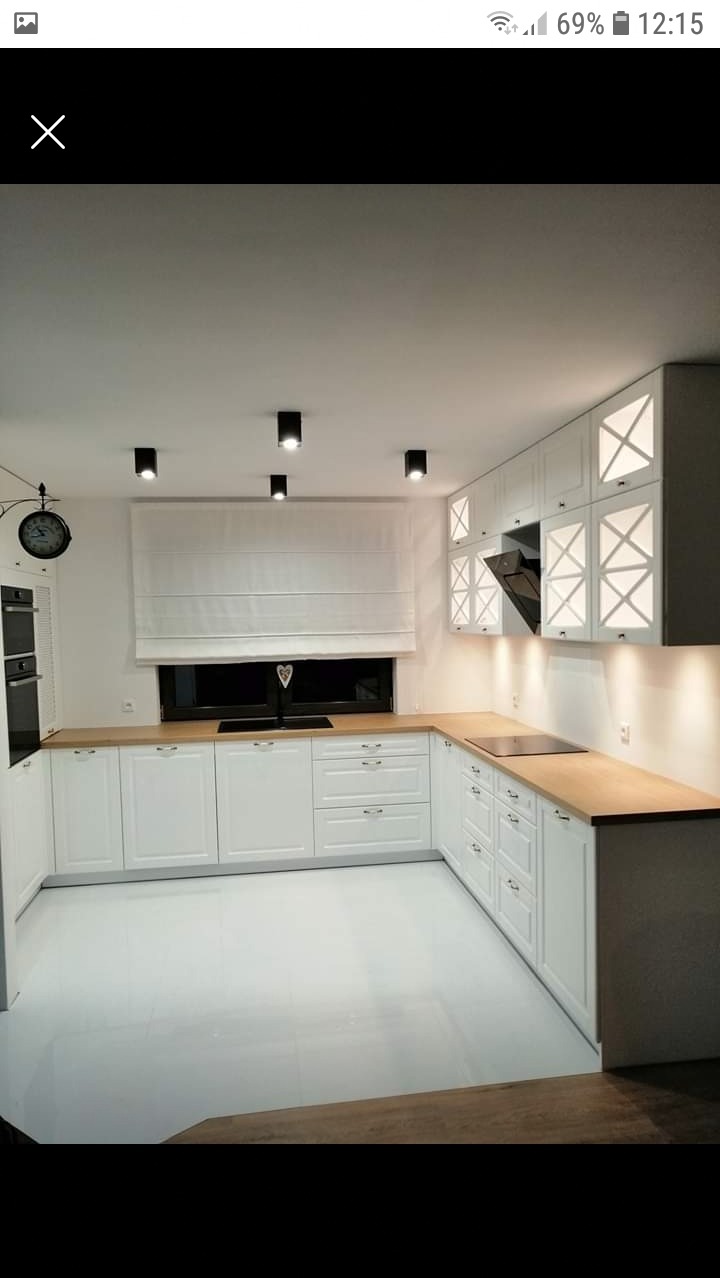 Nowoczesna, jasna kuchnia z białymi szafkami z drewnianymi blatami, czarnymi lampami sufitowymi i zegarem na ścianie.