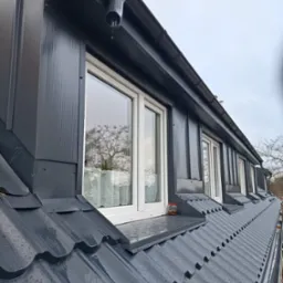 Szary dach z oknami dachowymi i metalowymi obróbkami blacharskimi. Widoczne krople deszczu na dachu i rynna z odpływem. W oknach odbija się drzewo.