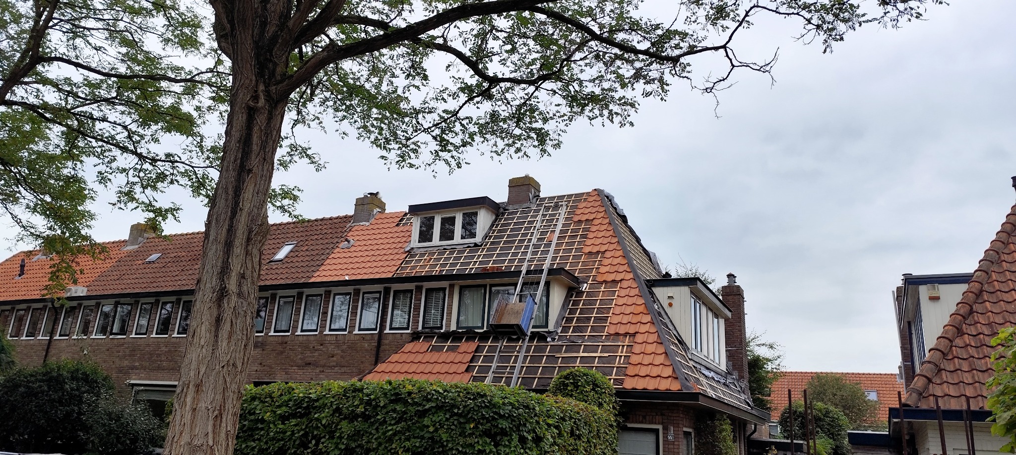 Częściowo zdjęty dach z czerwonej dachówki na dwupiętrowym domu. Widoczne łaty, drabina i fragmenty dachówek. Prace dekarskie w toku, pochmurne niebo w tle.