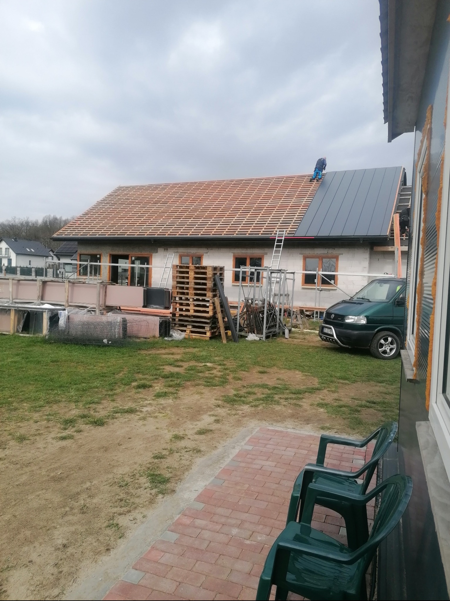 Montaż dachu z blachy na budynku jednorodzinnym. Widoczne rusztowanie, palety z materiałami i częściowo ułożona dachówka. Prace dekarskie w toku, pochmurne niebo.