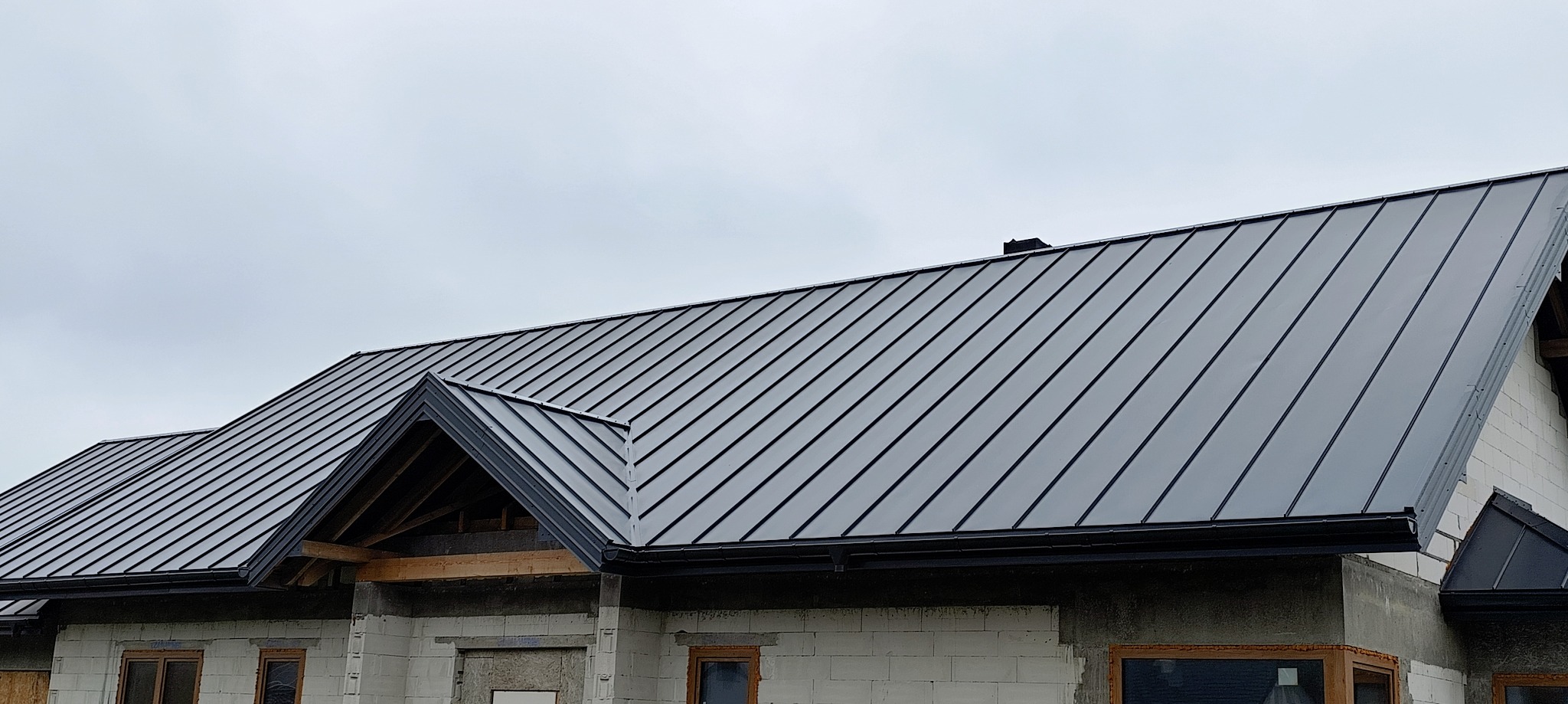Nowoczesny, grafitowy dach z blachy na surowym budynku. Widoczne obróbki blacharskie i rynny. Ujęcie z dołu, pochmurne niebo w tle.