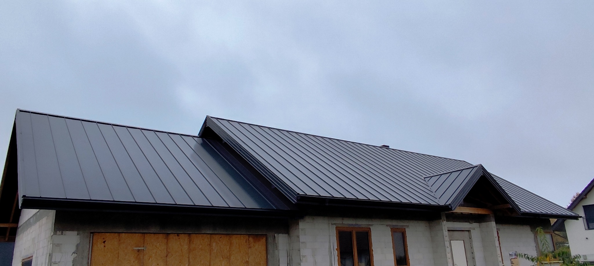 Nowy, grafitowy dach z blachy na surowym budynku w trakcie budowy, widoczny na tle pochmurnego nieba. Elementy obróbki blacharskiej podkreślają nowoczesny wygląd.