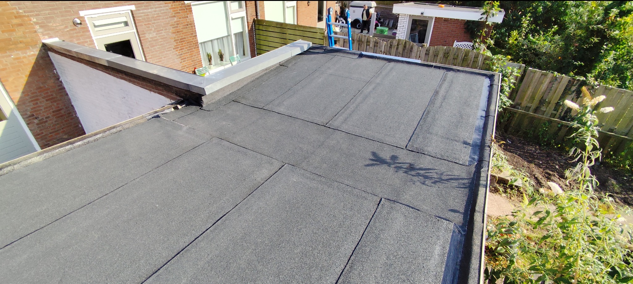 Wykończony, płaski dach pokryty papą bitumiczną, z widocznymi łączeniami i obróbką blacharską. W tle budynek z oknami i zielenią.