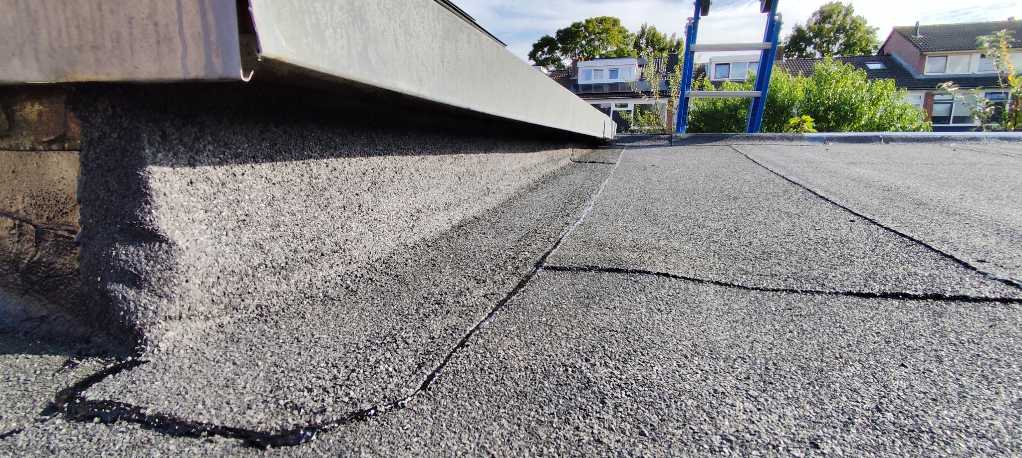 Szczegółowe ujęcie płaskiego dachu pokrytego papą z widoczną obróbką blacharską i drabiną w tle, ukazujące detale wykonania i wykończenia.