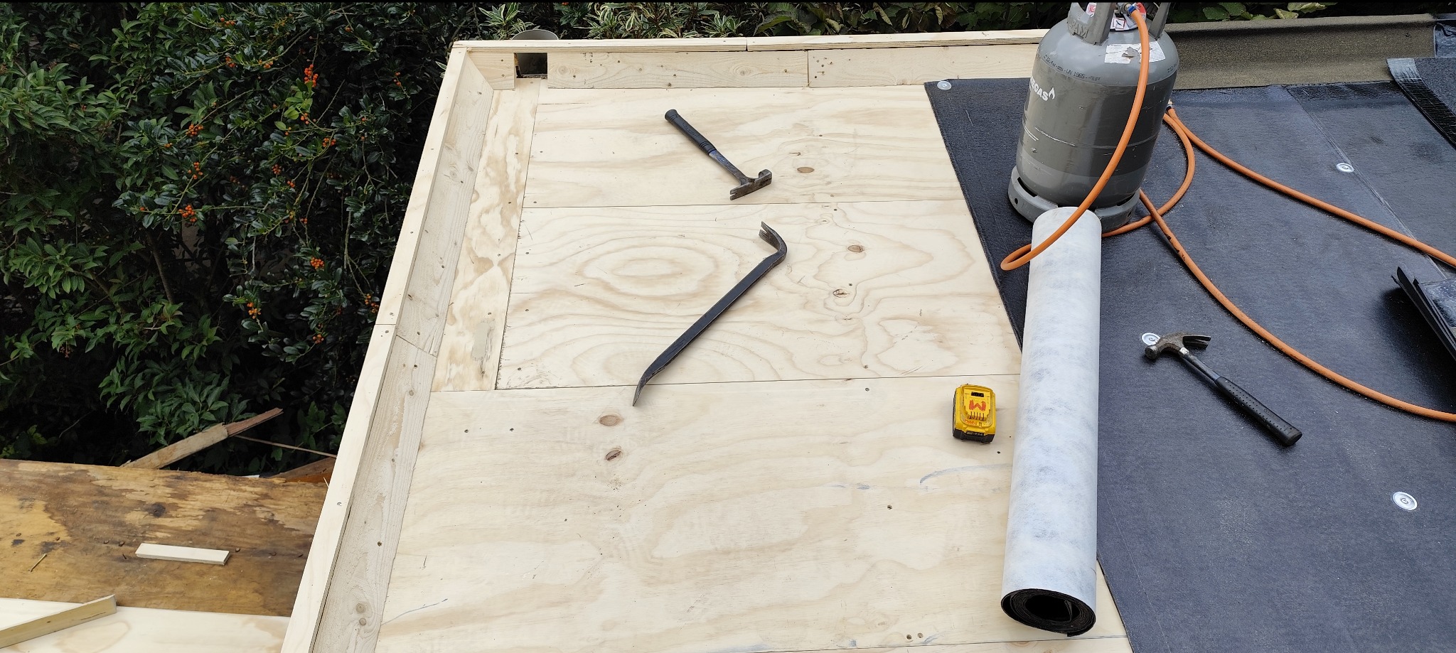 Etap budowy dachu płaskiego z deskowaniem, narzędziami i papą. Widoczna butla gazowa, młotki i łom. Drzewa w tle.