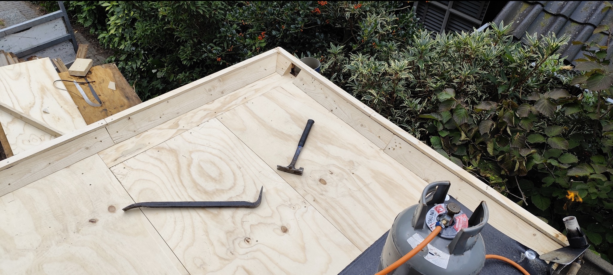 Konstrukcja dachu z płyt OSB, narzędzia dekarskie (młotek, łom) i butla gazowa na tle zieleni. Widok z góry na etap budowy dachu.