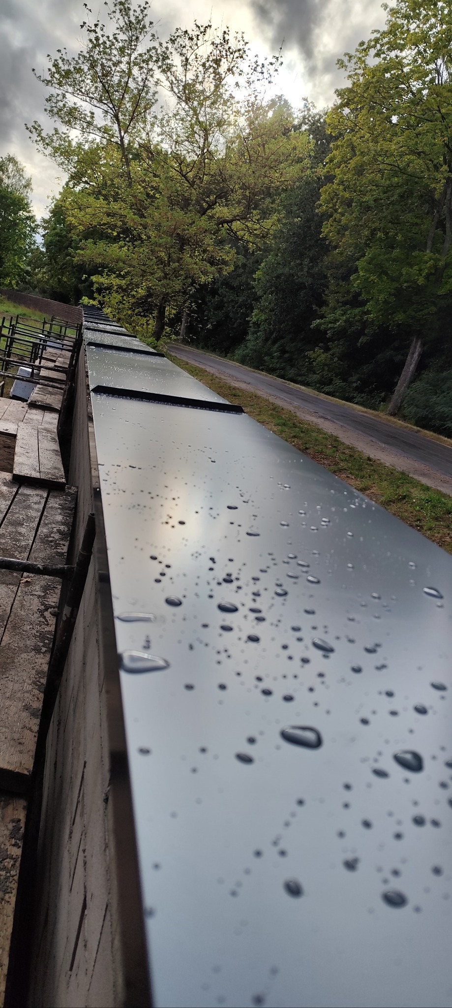 Błyszcząca, metalowa obróbka blacharska dachu z kroplami wody, widok z góry, w tle zieleń drzew i fragment drogi, pochmurne niebo.