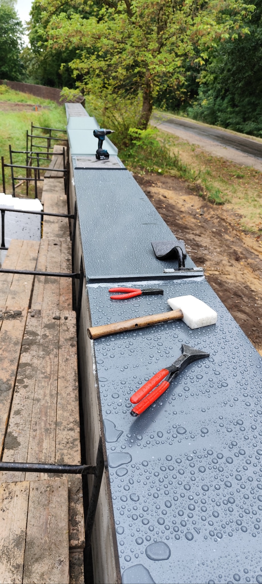 Mokry, szary parapet z narzędziami dekarskimi: młotek, kombinerki, wiertarka. W tle zieleń, rusztowanie. Widok z góry na placu budowy, po deszczu.