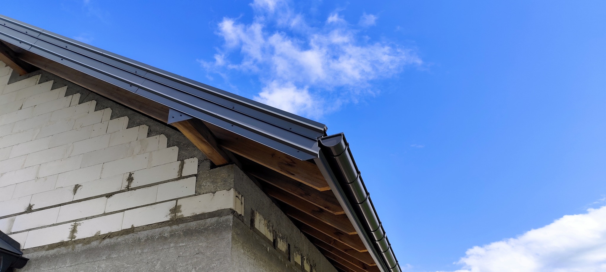 Fragment dachu z czarną blachą i rynną, widoczny mur z bloczków betonowych pod błękitnym niebem z chmurami. Ujęcie z dołu, podkreślające detale wykończenia.