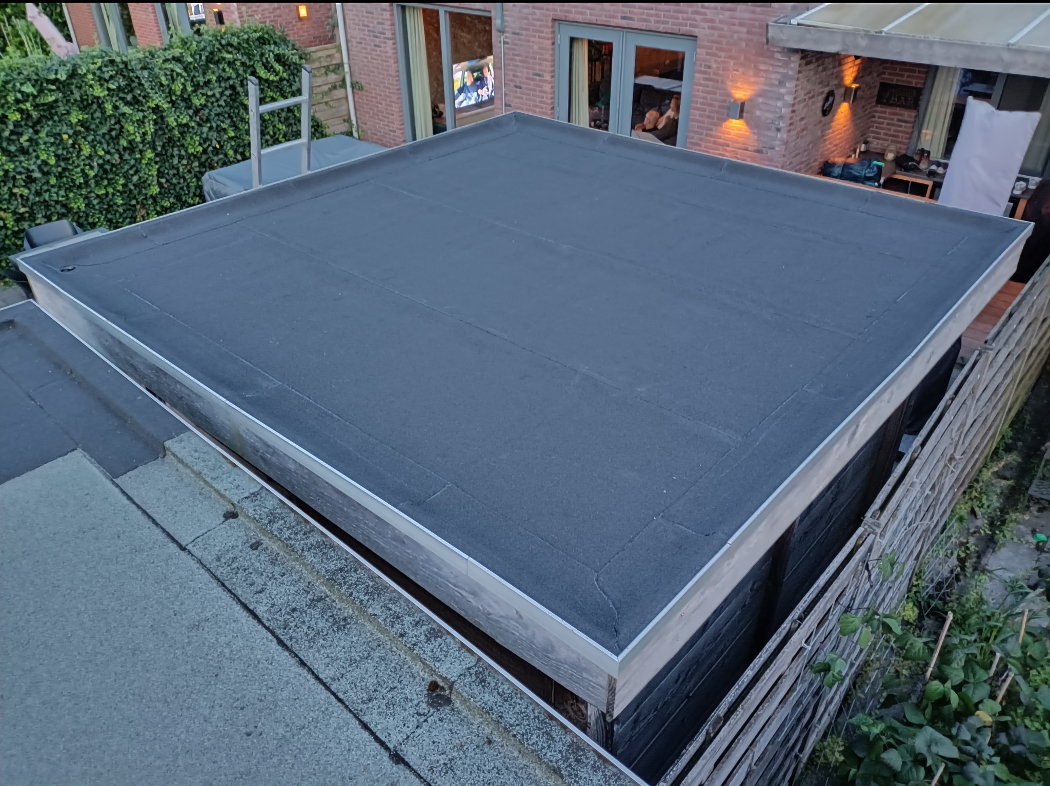 Wykończony, płaski dach z bitumiczną membraną, obróbkami blacharskimi i drewnianą konstrukcją. Widok z góry na tle zieleni i zabudowań.