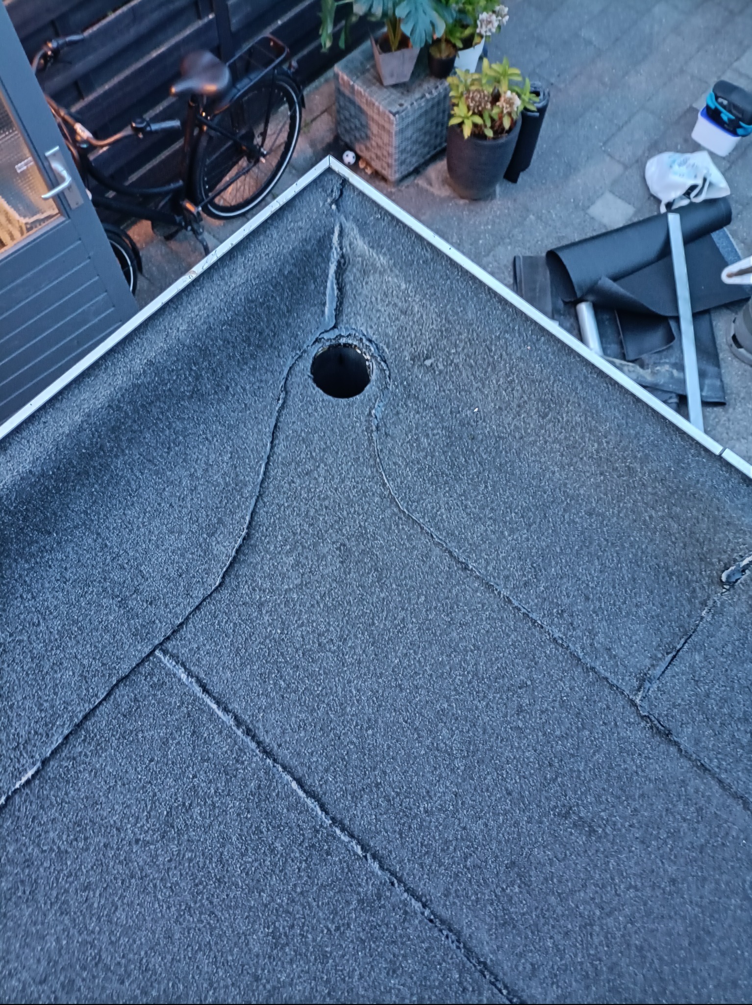 Detale pokrycia dachu papą z widocznym otworem odpływowym, materiały budowlane i rower w tle. Układanie papy na dachu płaskim.