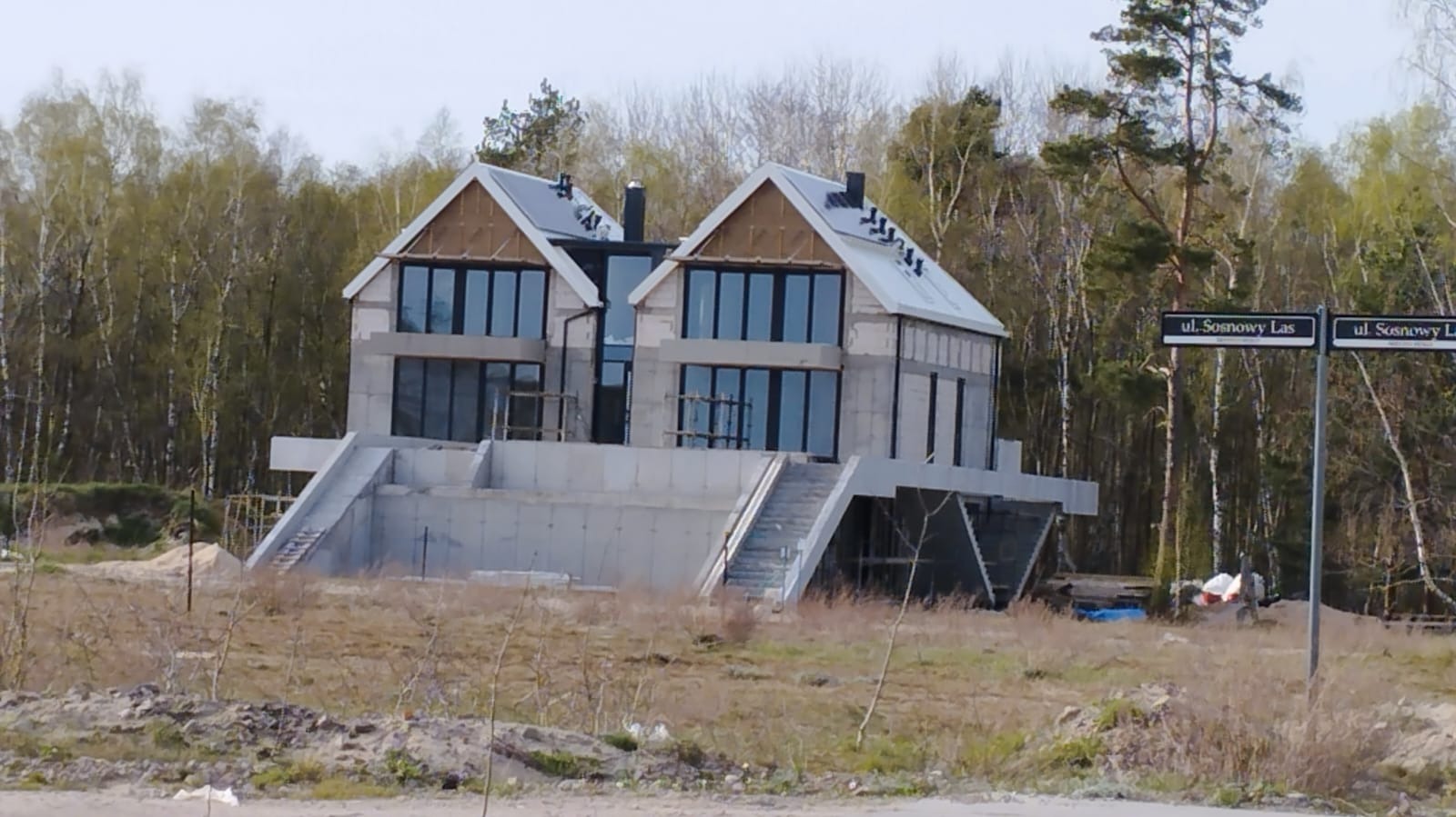 Surowy stan dwukondygnacyjnego domu z dużymi oknami, betonowym fundamentem i schodami. Widoczne nieocieplone poddasze i tabliczka z nazwą ulicy 'ul. Sosnowy Las'.