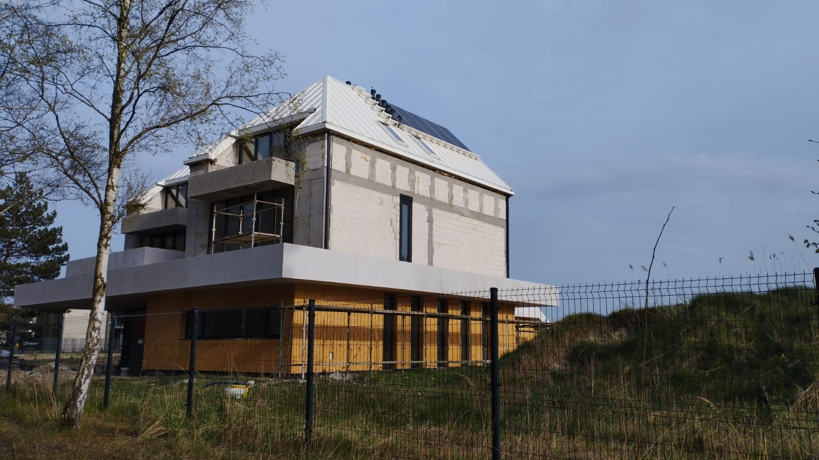 Nowoczesny dom w trakcie budowy: biała blacha na dachu, panele słoneczne, surowy beton, drewniane elementy. Widok zza ogrodzenia.