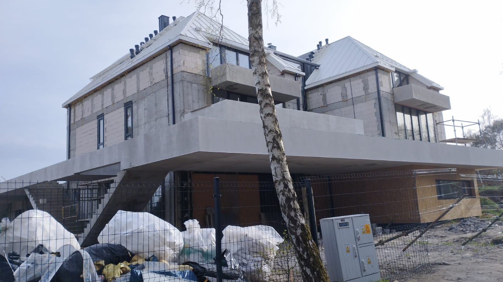 Dom w trakcie budowy z elementami wykończeniowymi dachu, widoczne białe bloczki i metalowa konstrukcja, częściowo zasłonięty przez brzozę i worki z odpadami.