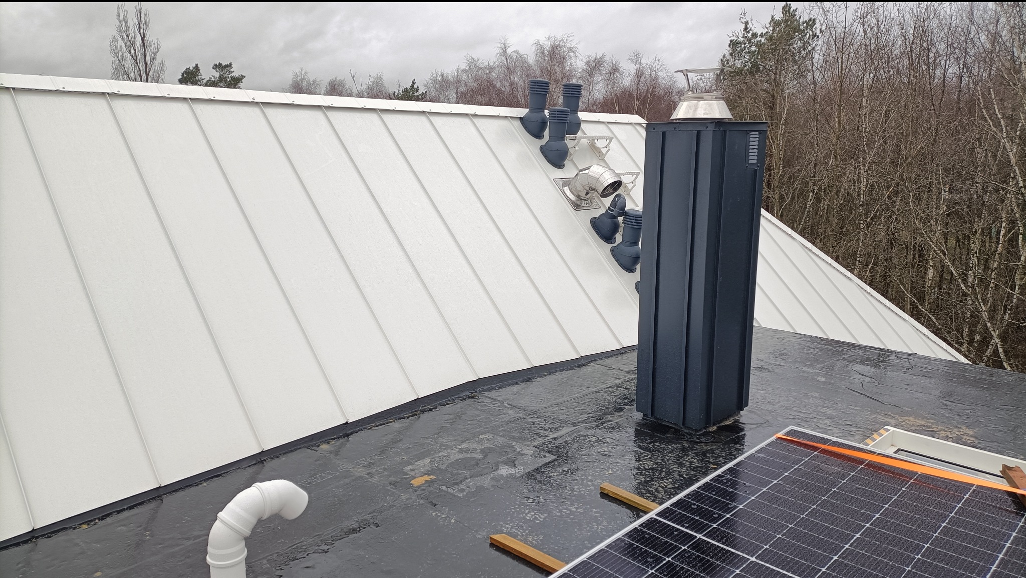 Biały dach panelowy z kominem i wentylacją, widok z góry na mokrą, czarną papę z panelem słonecznym w rogu, zimowy krajobraz w tle.