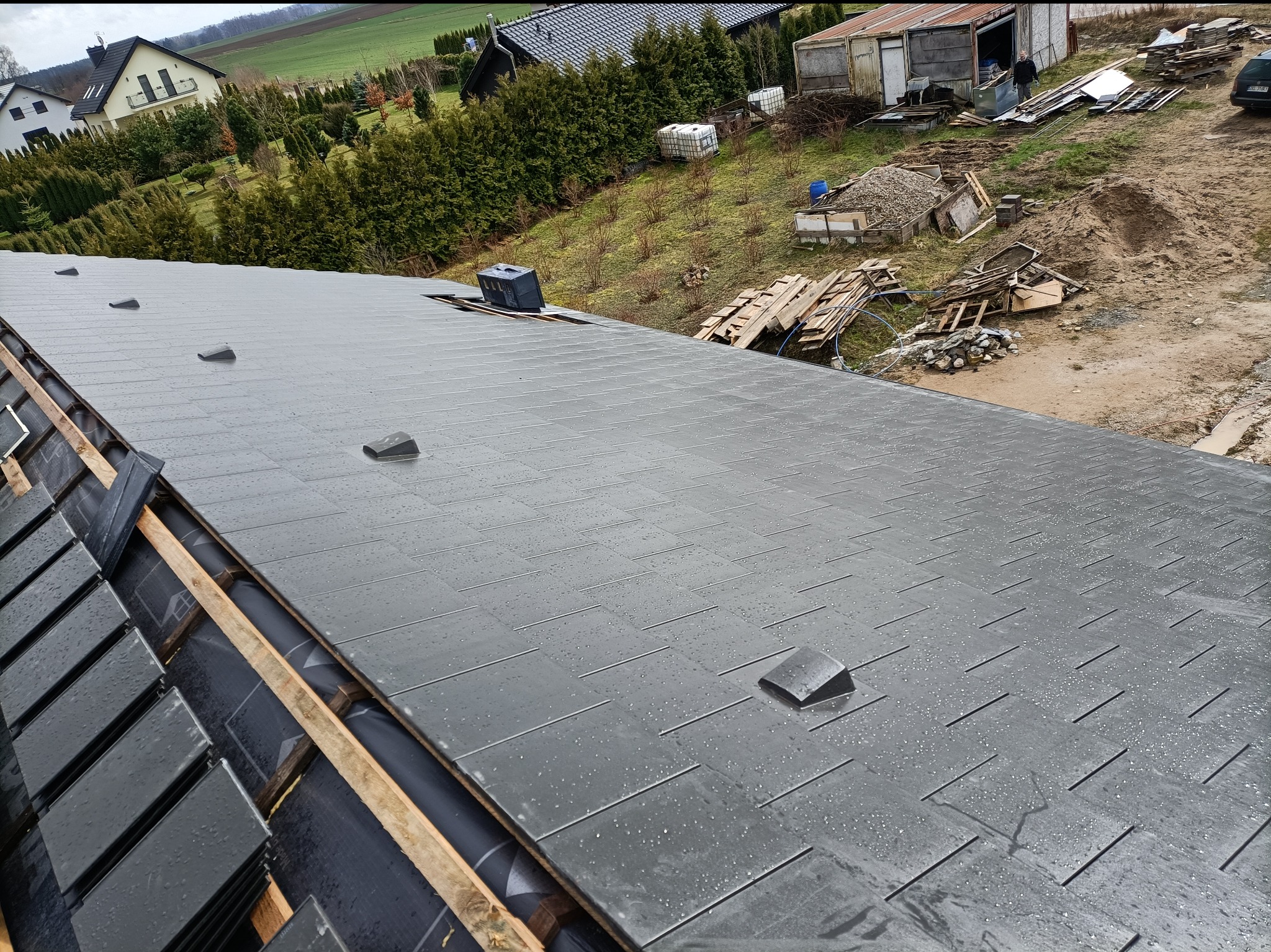 Nowy, grafitowy dach z dachówki ceramicznej ułożony w karo. Widoczne elementy konstrukcji i detale wykończeniowe. Dach jest mokry od deszczu.