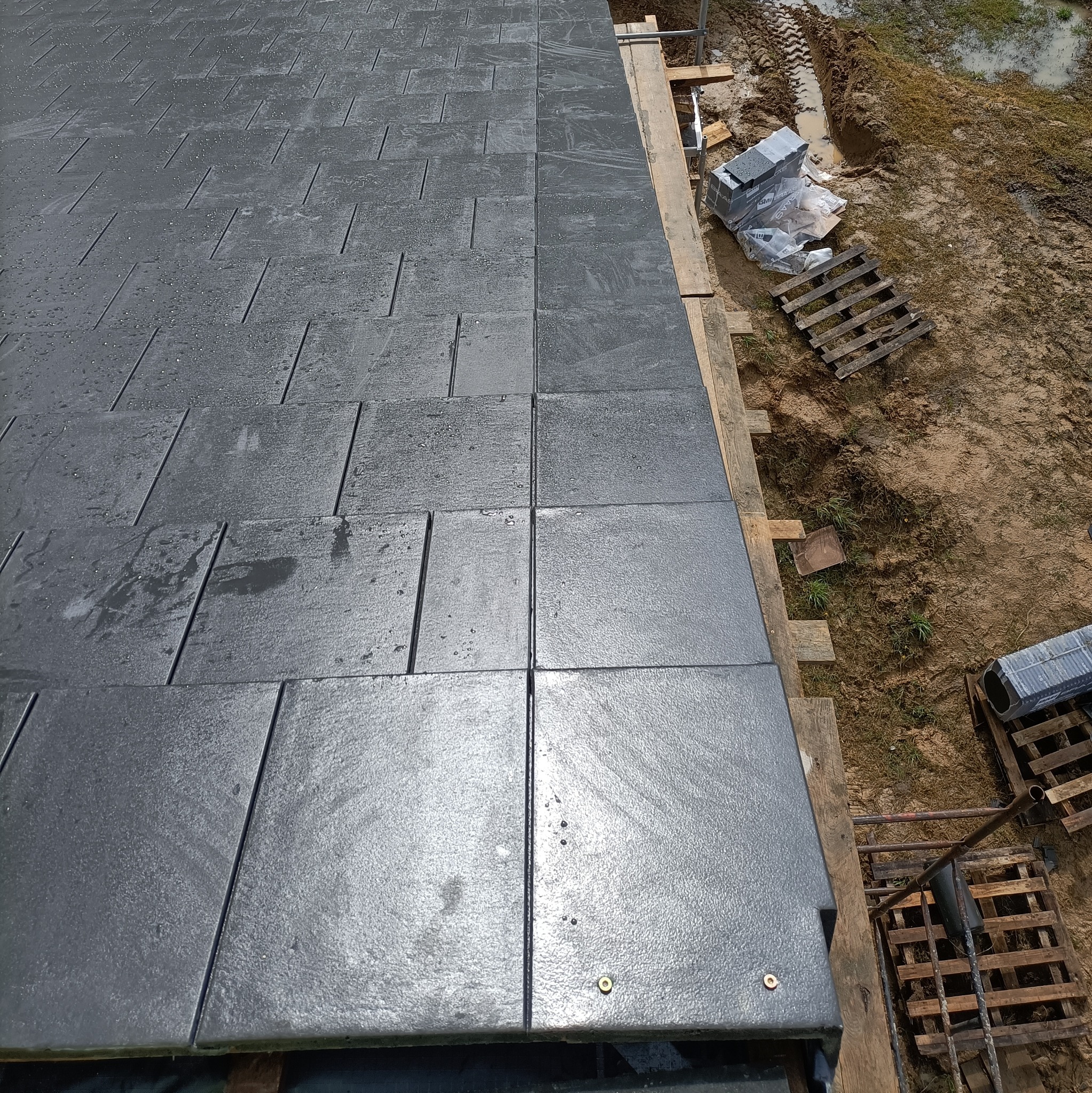 Nowy, grafitowy dach z dachówki betonowej na mokro, z widocznymi elementami rusztowania i zapasem materiału na błotnistym podłożu, wykonany z dbałością o detale.