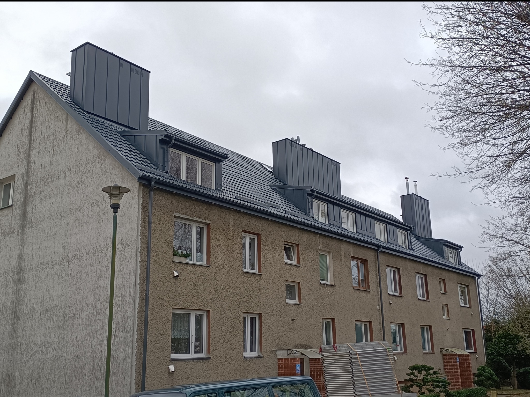 Budynek wielorodzinny po renowacji dachu z blachy w kolorze grafitowym. Widoczne kominy również obłożone blachą. Na pierwszym planie lampa uliczna i fragment rusztowania.
