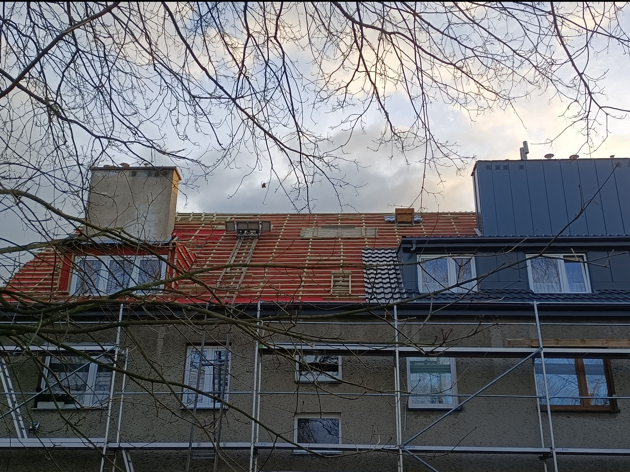 Budynek w rusztowaniach z częściowo zdjętym dachem. Widoczne elementy konstrukcji dachu i fragmenty nowego pokrycia. Gałęzie drzew na pierwszym planie.