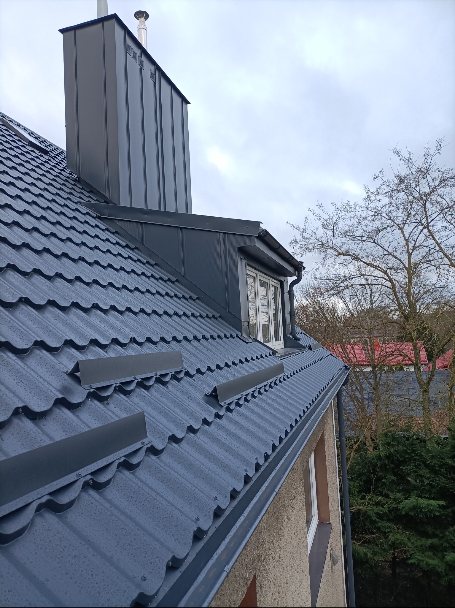 Szary dach z blachy trapezowej z kominem i oknem dachowym, widok z dołu, po deszczu. Na tle drzewa bez liści i pochmurne niebo.