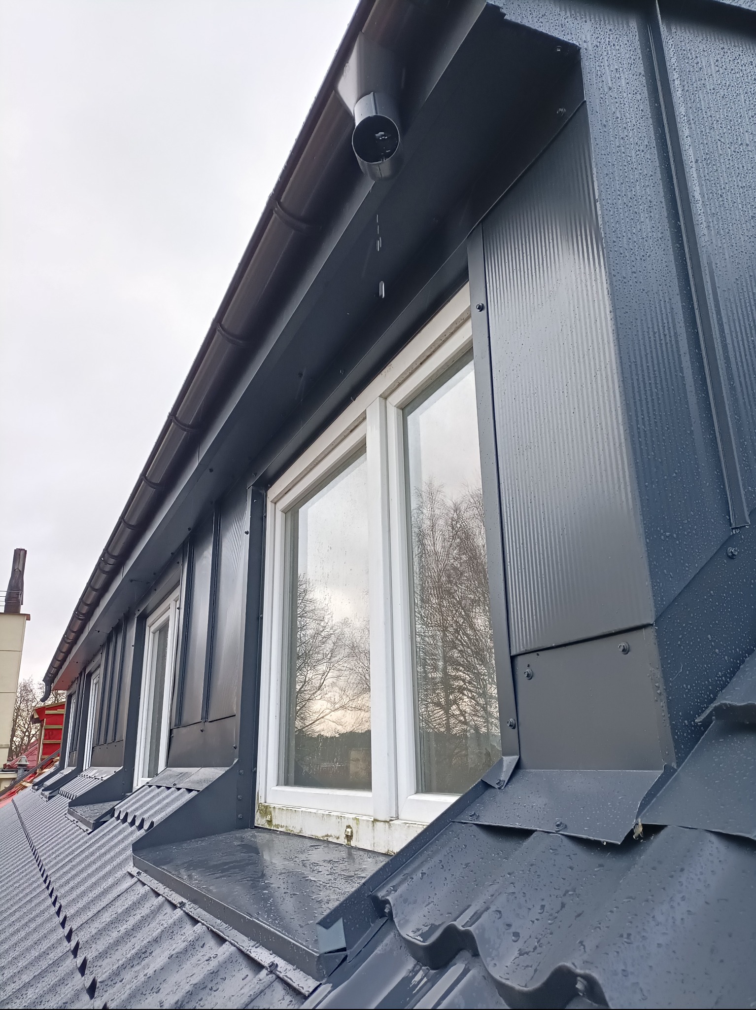 Szare, metalowe obróbki blacharskie okien dachowych z rynnami, po deszczu, widok z dołu pod kątem. Detal konstrukcyjny.