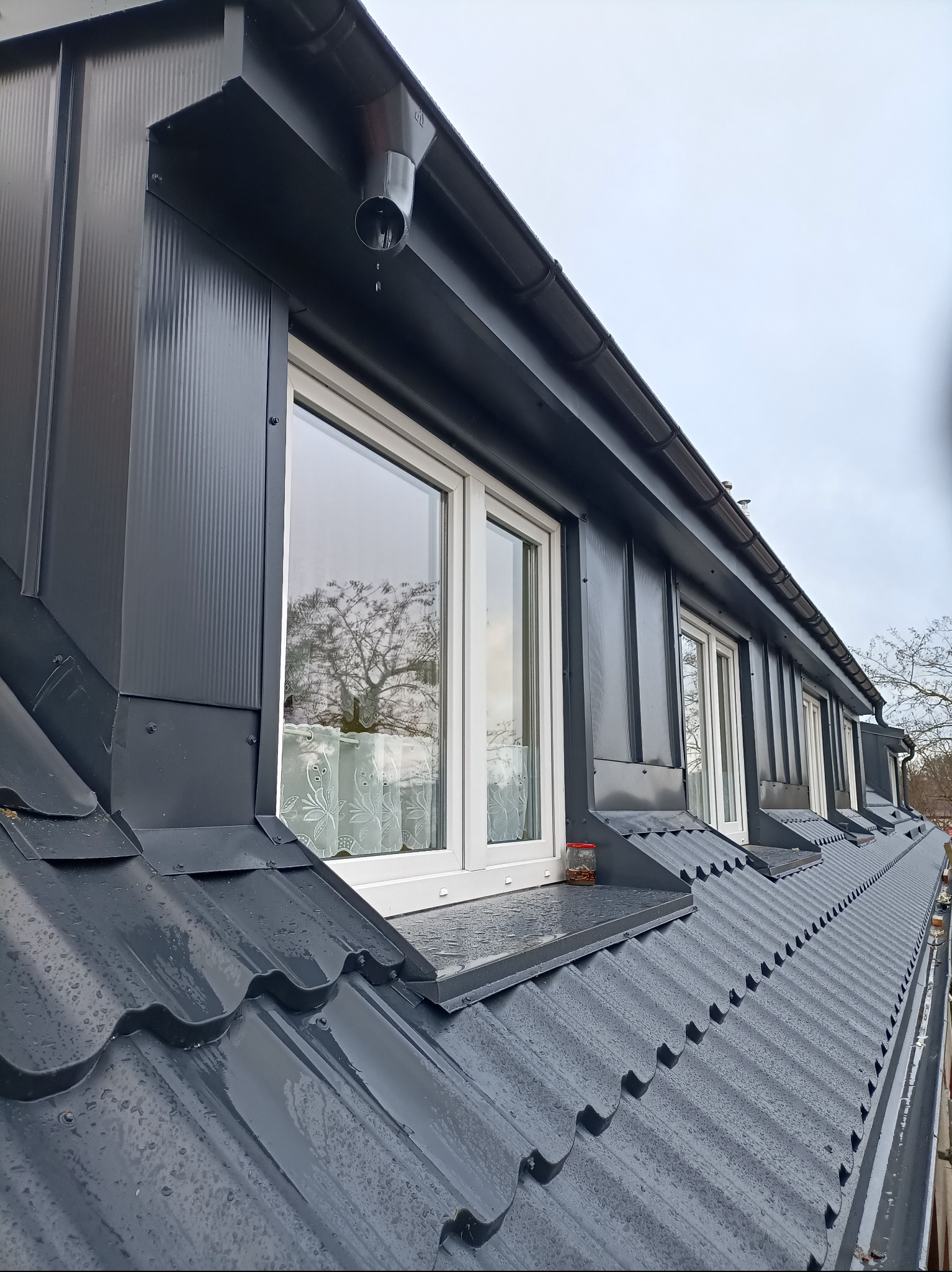 Szary dach z oknami dachowymi i metalowymi obróbkami blacharskimi. Widoczne krople deszczu na dachu i rynna z odpływem. W oknach odbija się drzewo.