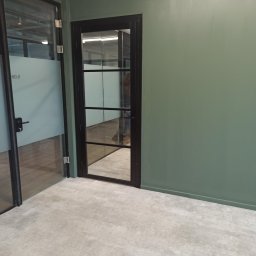 BUDEX - Wnętrze biurowe z pomalowaną na zielono ścianą, szklanymi drzwiami z czarną ramą i betonową podłogą. Widoczne elementy instalacji pod sufitem.