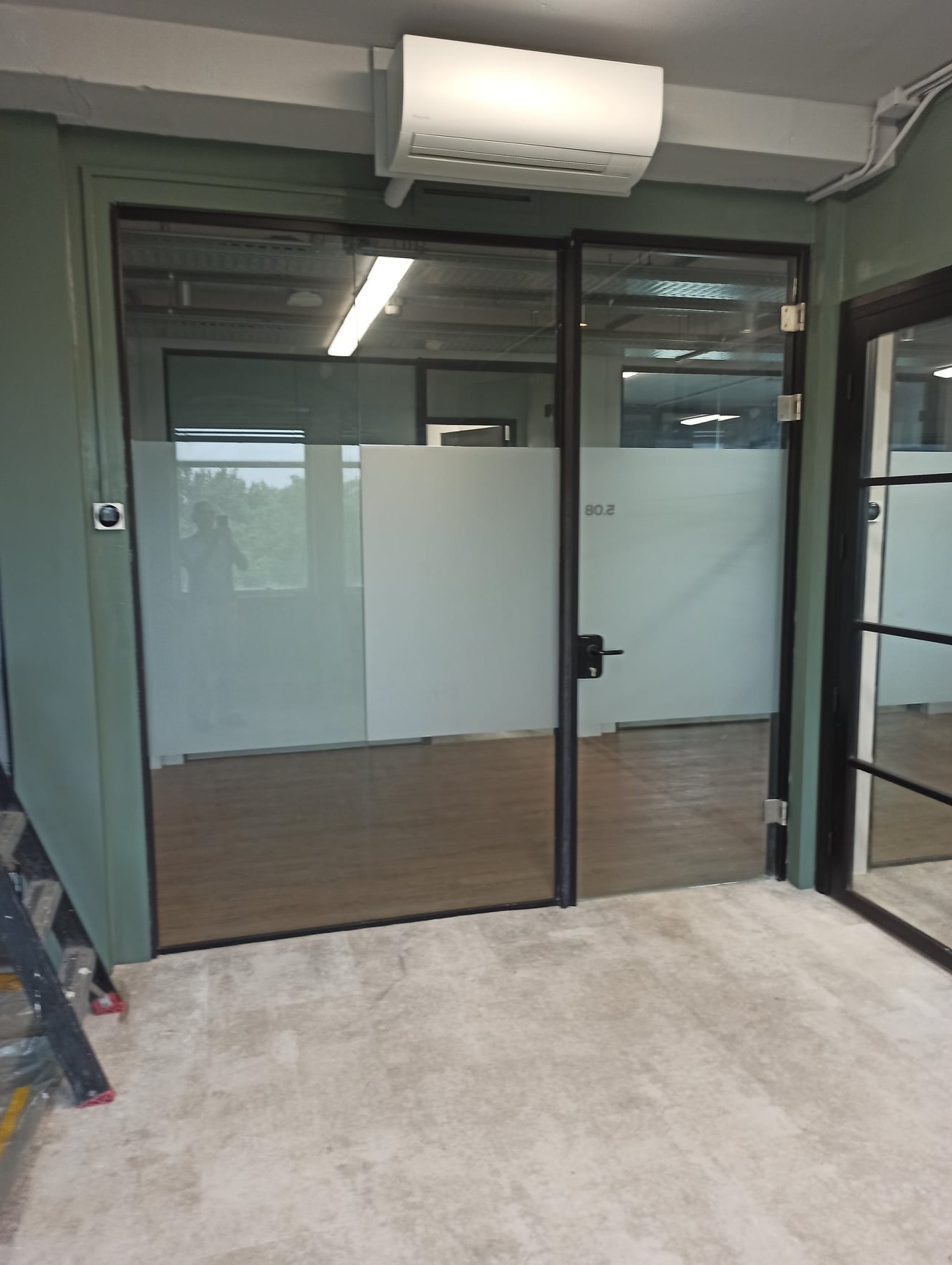 Biuro z przeszklonymi drzwiami z czarną ramą i matową folią na części szyby, klimatyzator na suficie, widoczne elementy rusztowania.