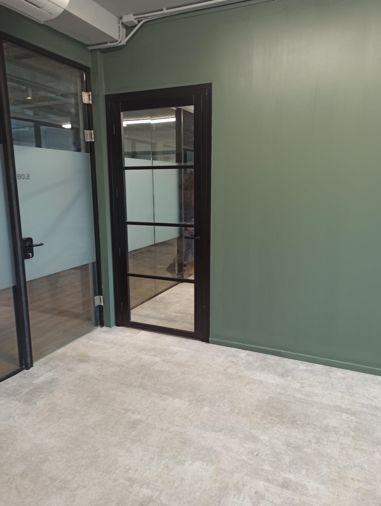 Wnętrze biurowe z zieloną ścianą, przeszklonymi drzwiami z czarną ramą i betonową podłogą. Widoczne elementy instalacji pod sufitem.